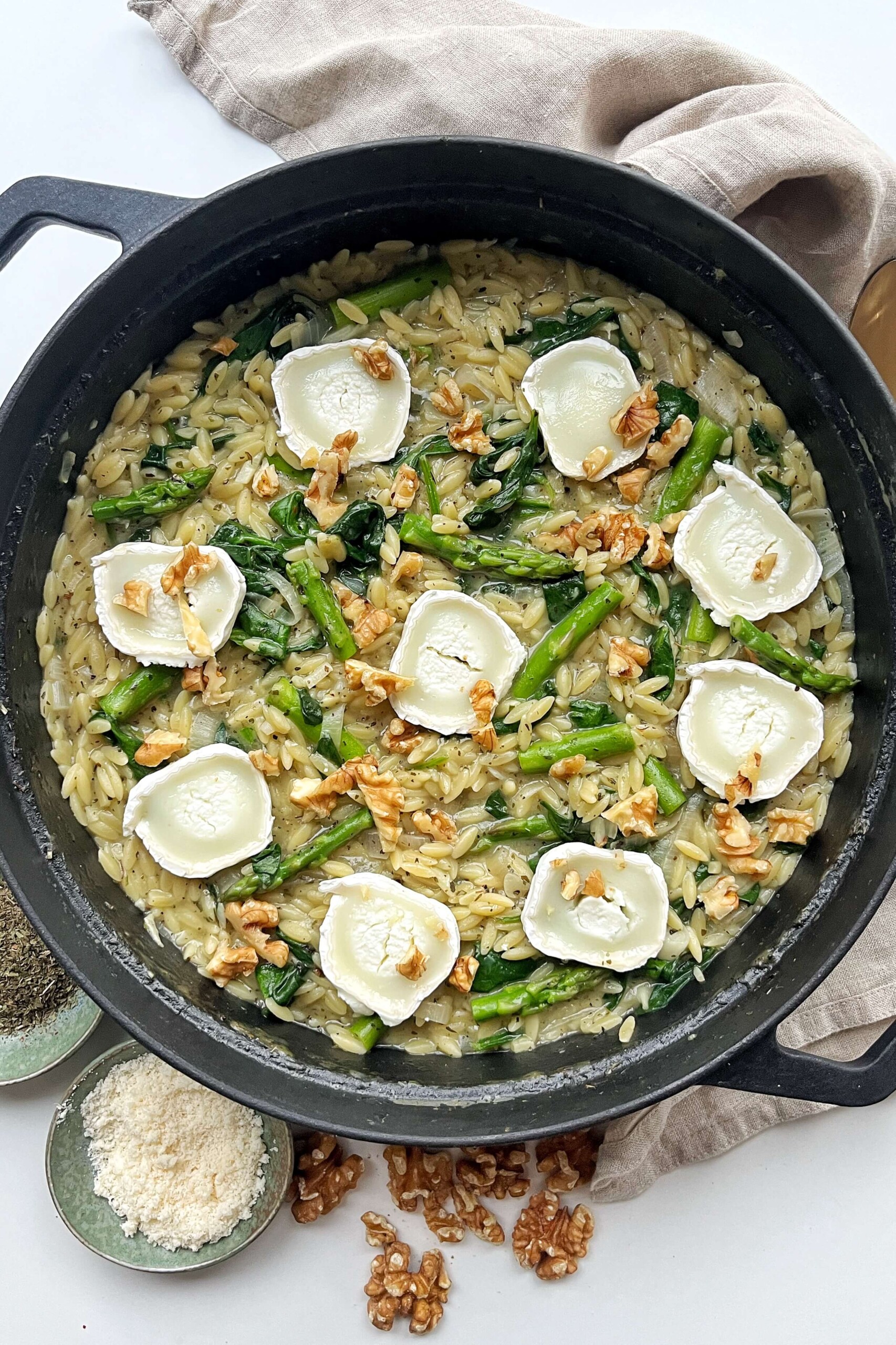 Asparagus orzotto topped with walnuts and goats cheese in a black pan.