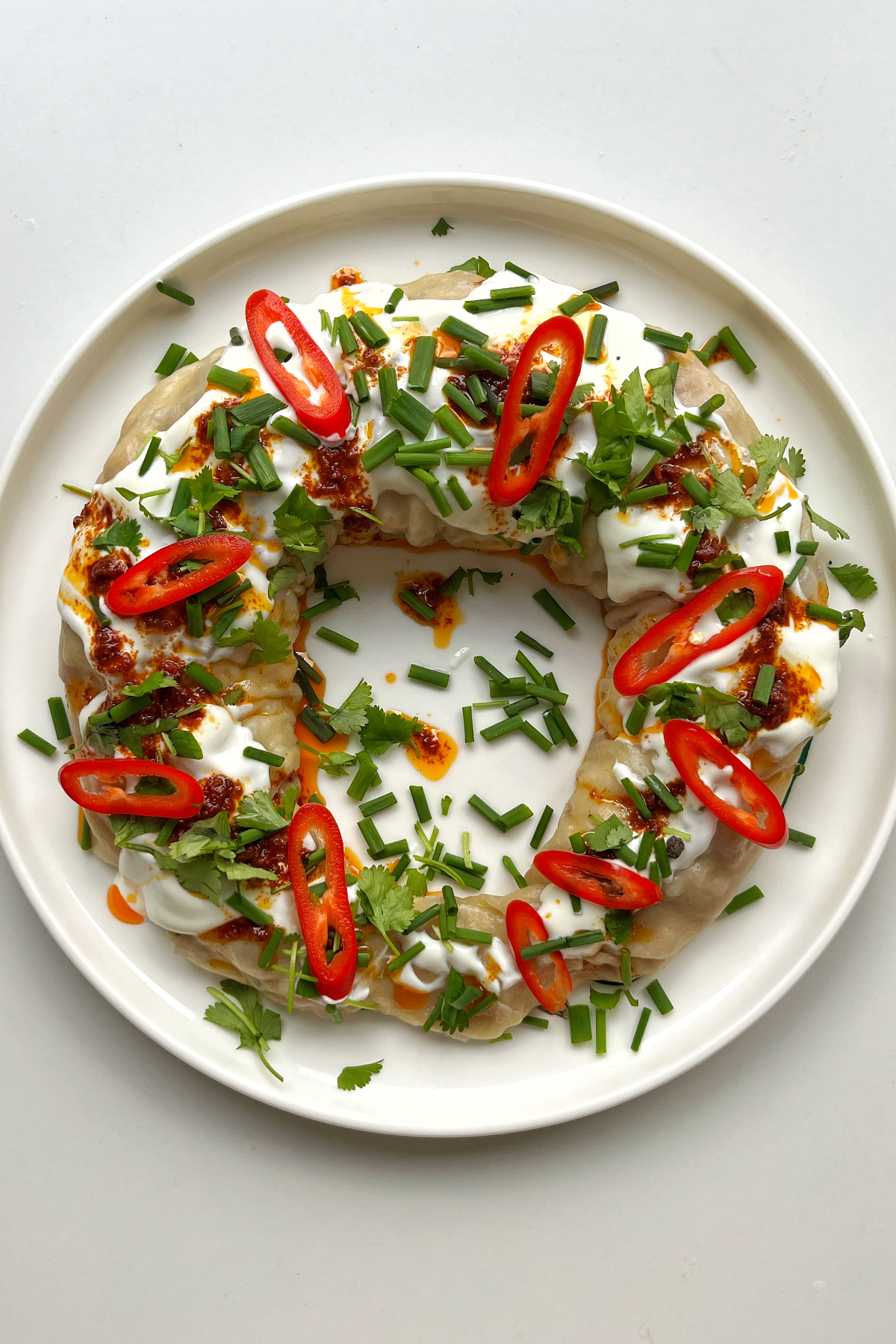 Dumpling wreath on a white plate topped with yoghurt, harissa oil, chives, and sliced chilli.
