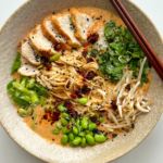Marry me chicken ramen in a ramen bowl with chopsticks topped with chicken, edamame beans, coriander, beansprouts, and spring onions.
