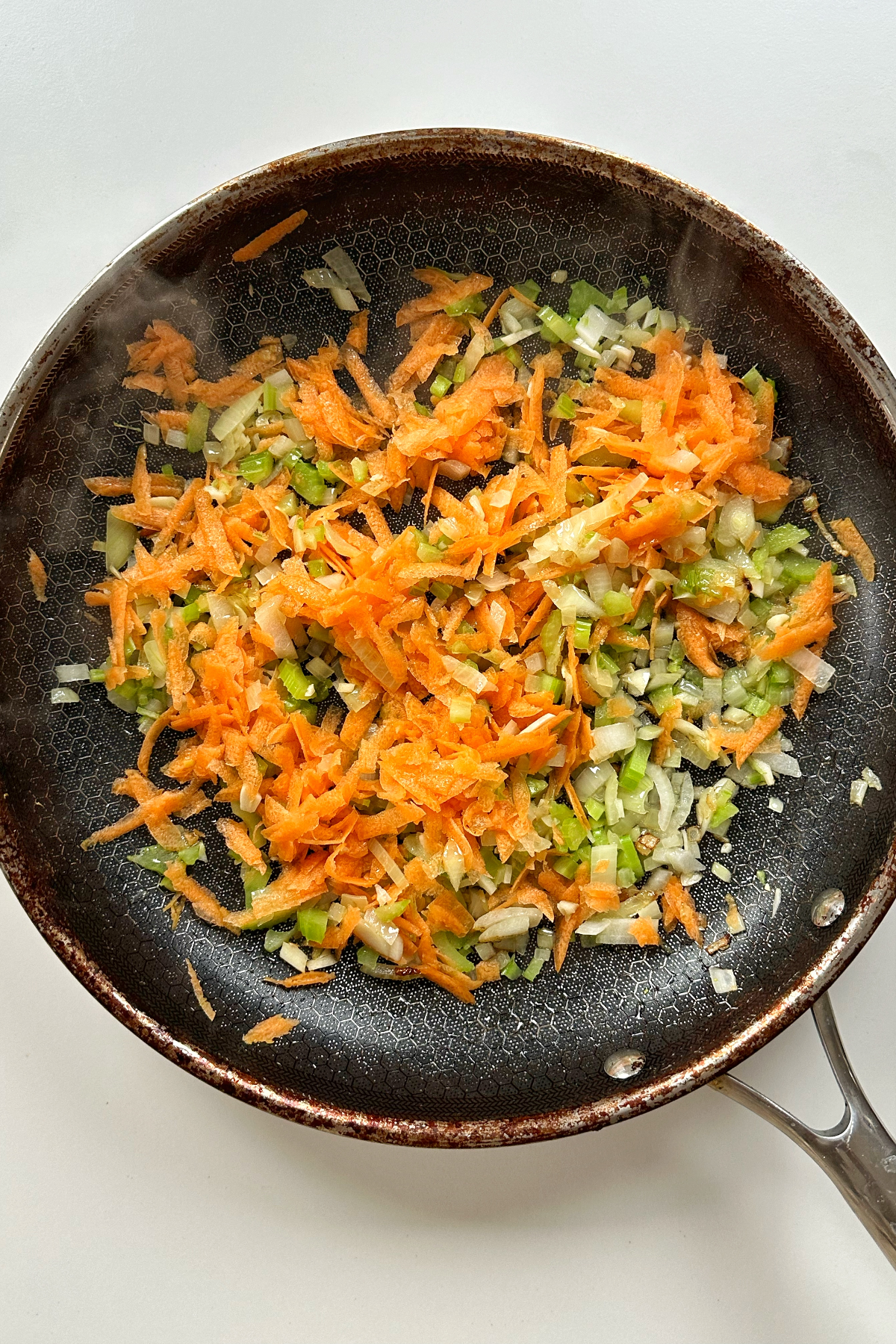 Frying off onion, garlic, celery, and carrot in a pan on a white background.