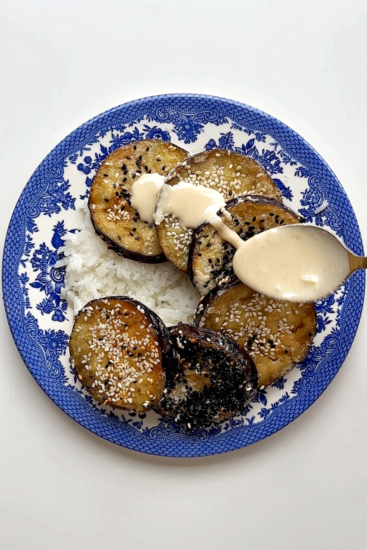 Drizzling maple tahini sauce over aubergines on a blue and white plate.