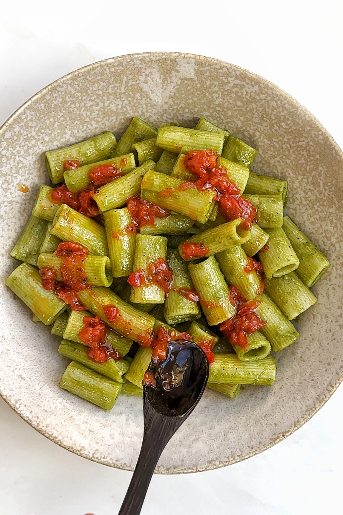 Drizzling tomato sauce over pasta using wooden spoon in cream bowl.
