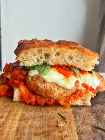 Sandwich with crispy eggplant, mozzarella, and marinara sauce on a chopping board.
