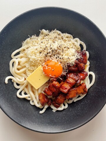 Black bowl with udon noodles, fried pork belly, grated parmesan, knob of butter, and egg yolk.