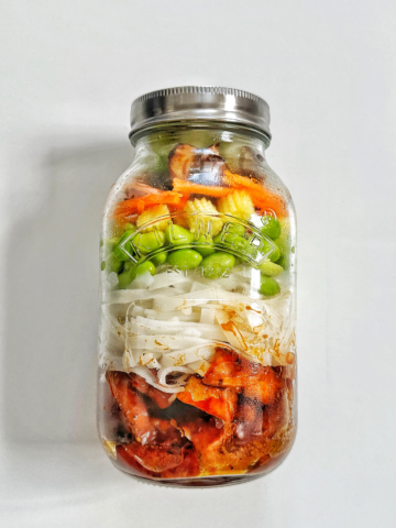 Jar of meal prep tofu with noodles and vegetables on a white background.