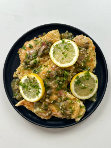 Chicken piccata on a black plate topped with lemon slices.