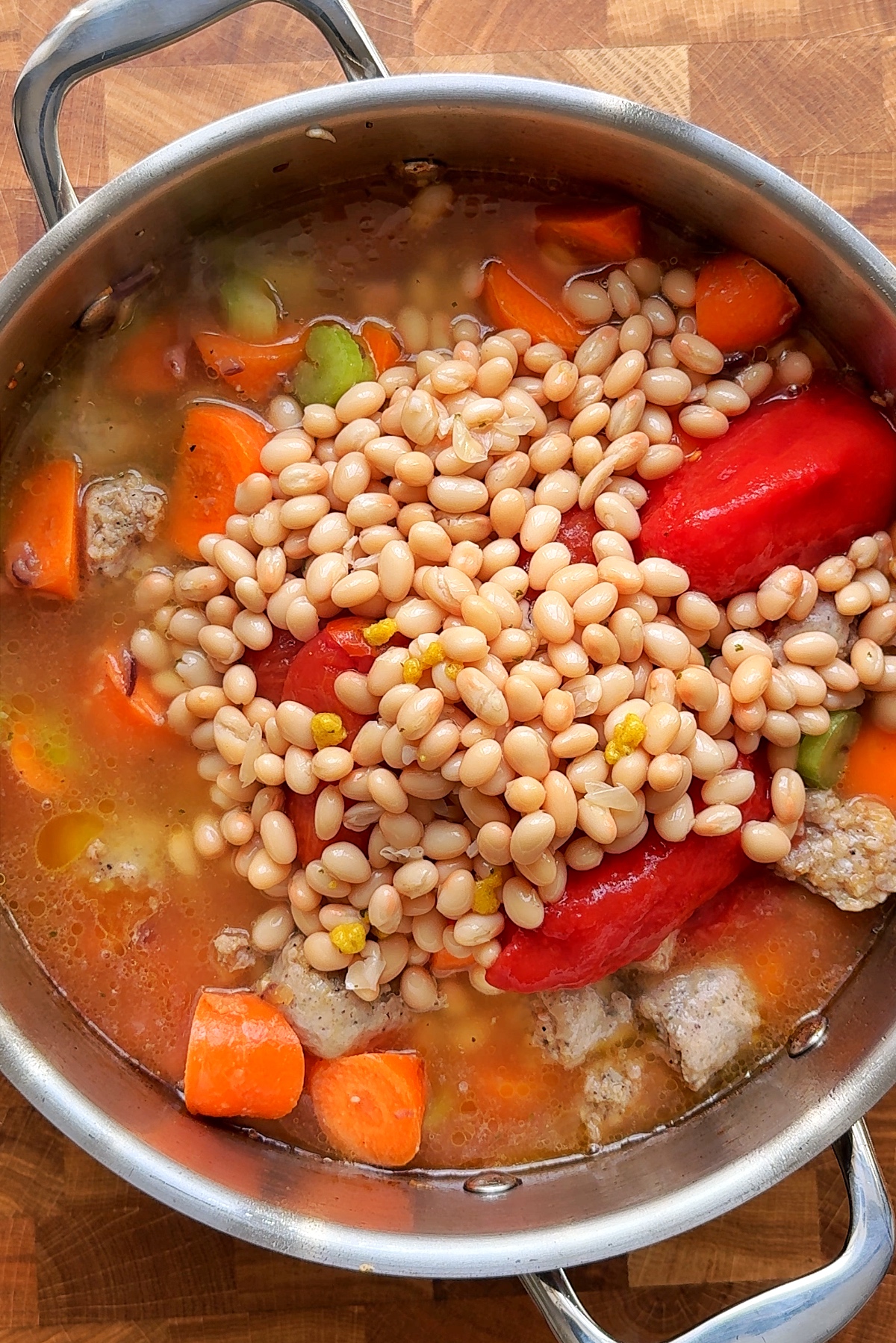Adding beans, tomatoes, carrots, celery to sausage stew.
