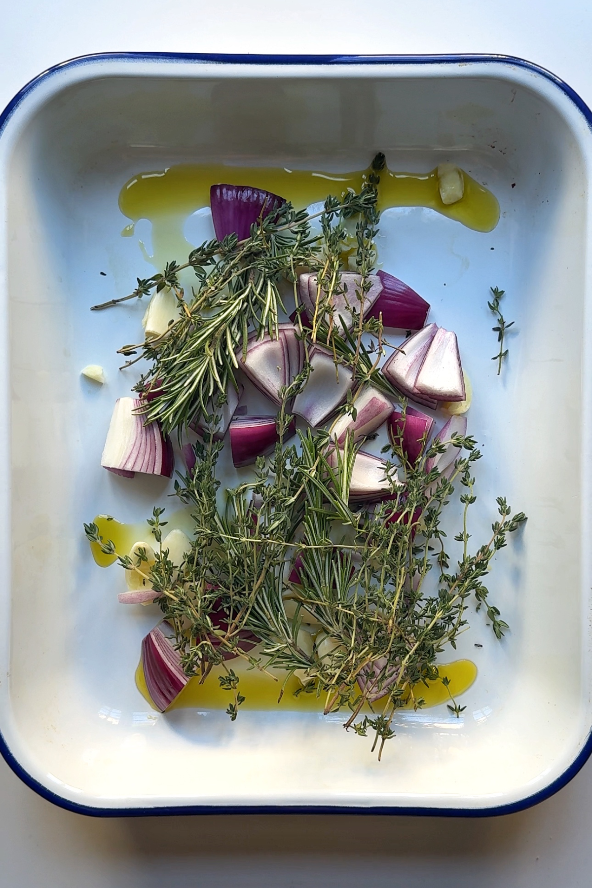 Red onion, garlic, thyme and rosemary in a tray with olive oil.