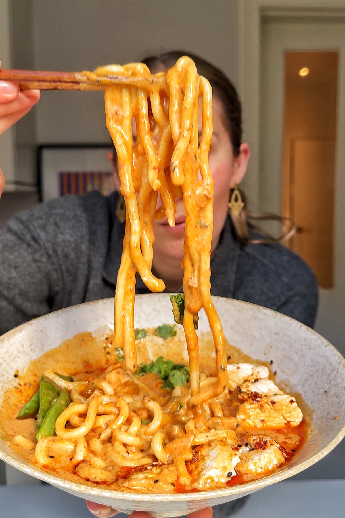 A woman pinching some peanut butter noodles with chopsticks and pulling them in front of her face.