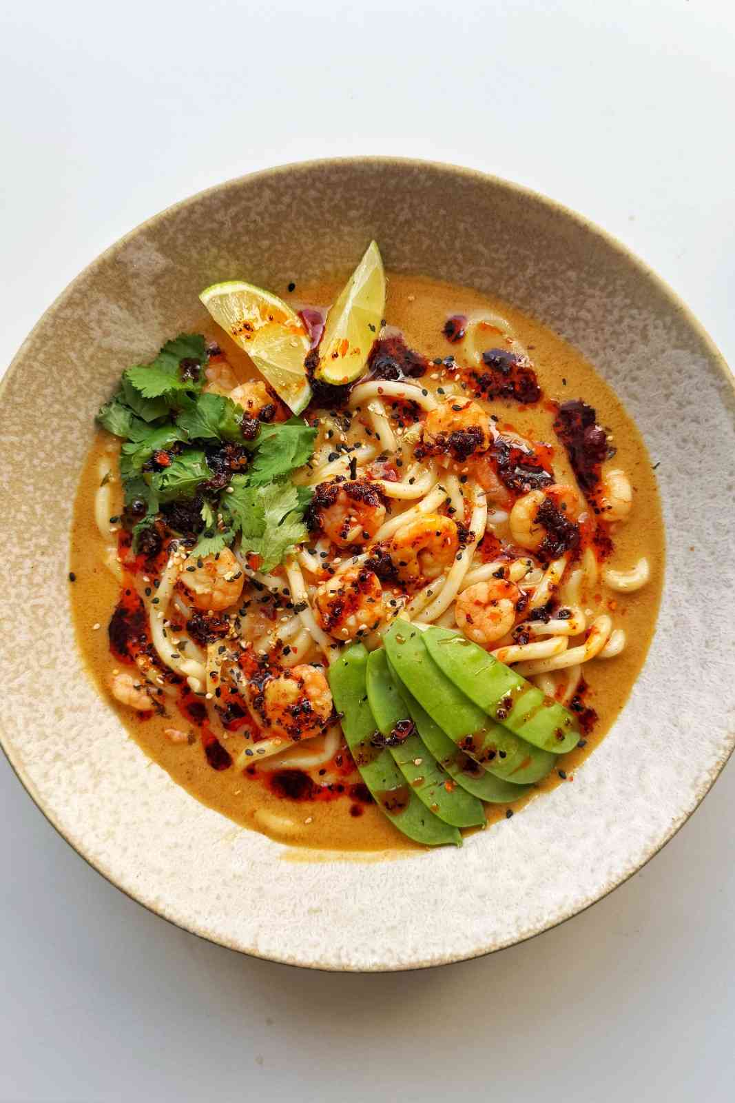 A bowl of creamy, orange-tinted ramen with small pink shrimps, bright green snow peas, fresh cilantro, and lime wedges, drizzled with spicy chili oil to showcase a speedy seafood protein hack.
