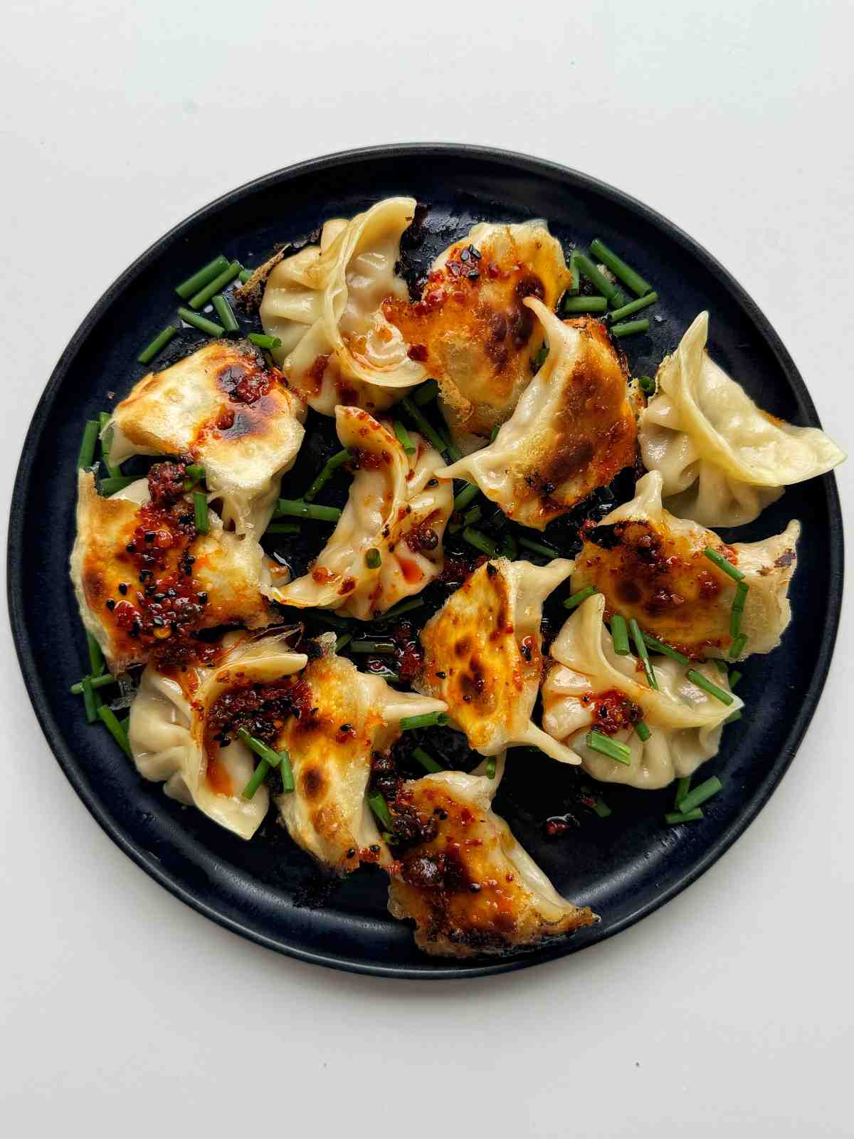 Pan-fried dumplings with crispy golden bottoms drizzled with chili oil and topped with fresh chives on a black plate