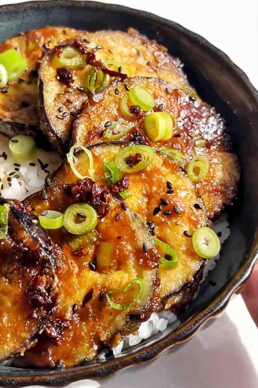 A black bowl filled with sticky miso eggplant and garnished with sesame seeds and spring onion.