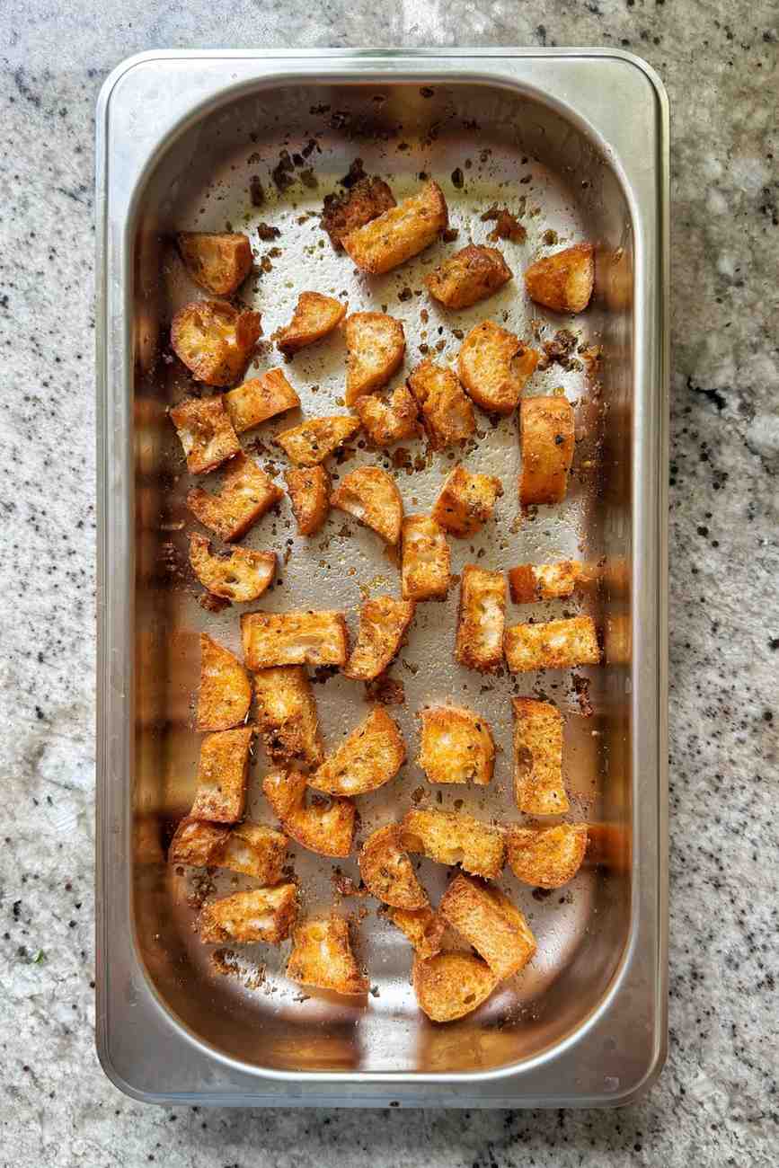 Seasoned, crunchy ciabatta croutons spread across a stainless steel pan after being toasted in a ninja air fryer.