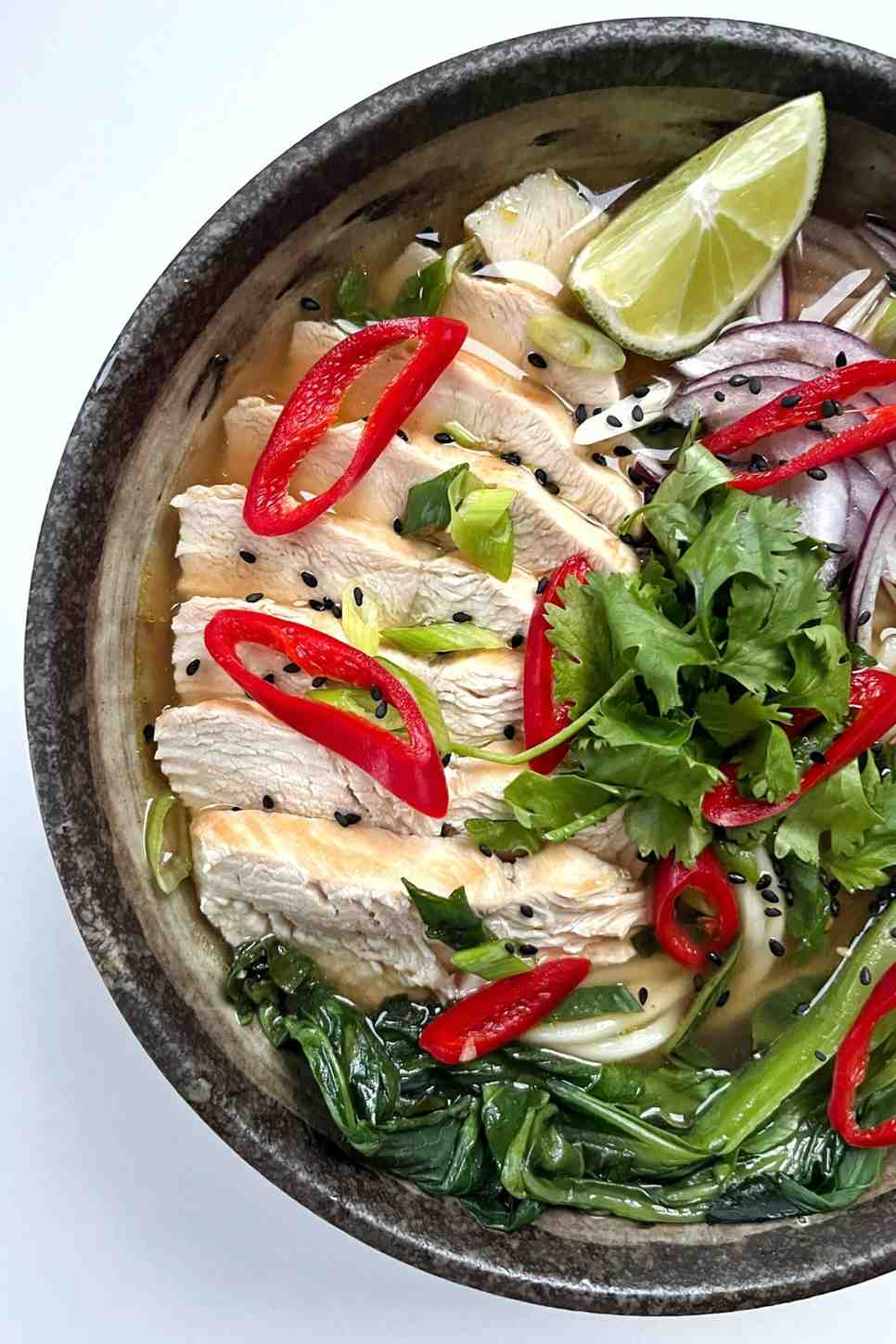 A bowl of chicken ramen noodles with pak choi, chili, red onion and lime as garnishes.