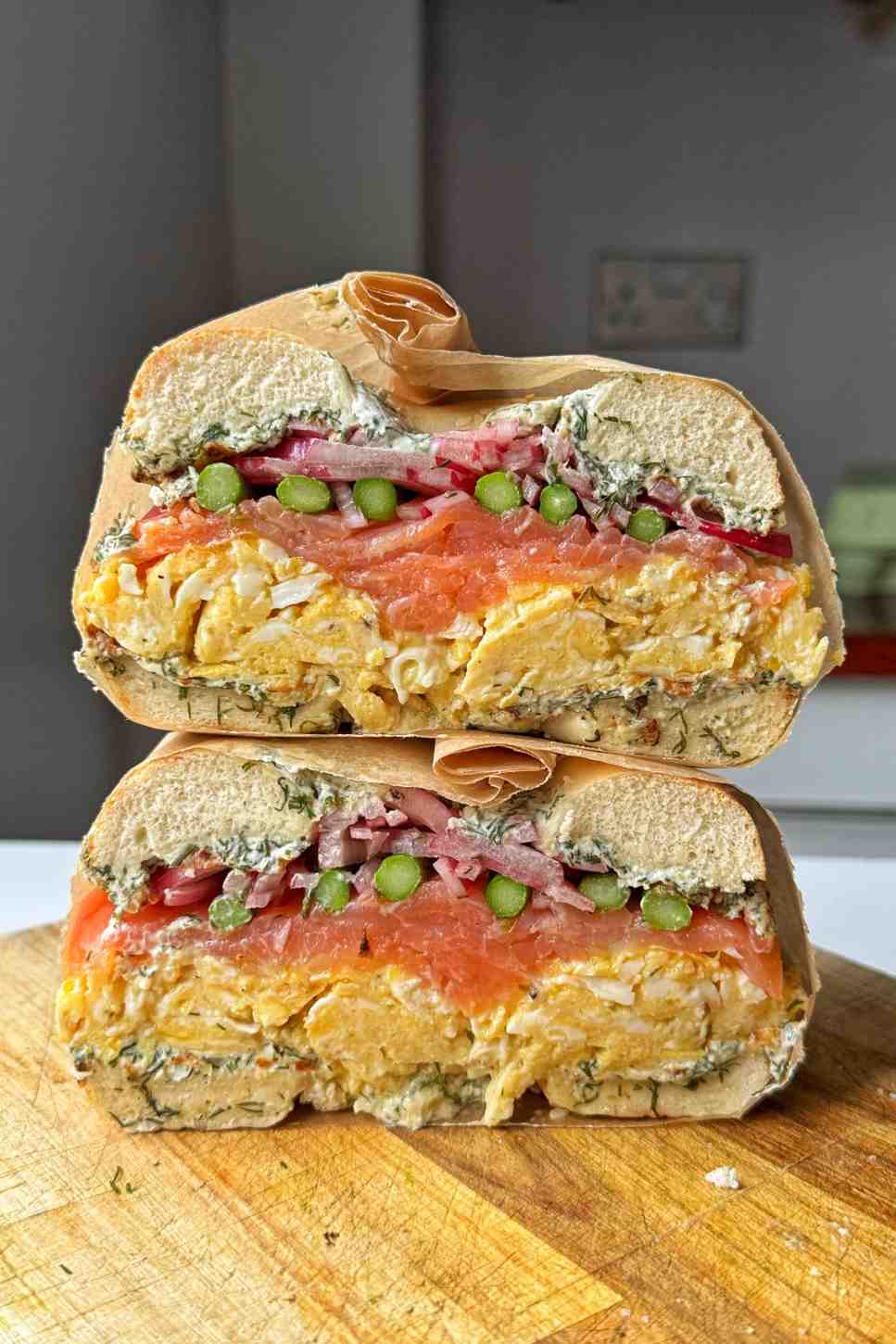Stacked smoked salmon bagel halves showing layers of dill cream cheese, scrambled eggs, smoked salmon, pickled red onions, and asparagus on a wooden cutting board