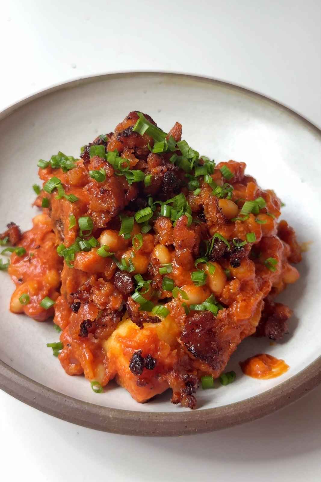 A loaded jacket potato topped with cheesy baked beans, crispy bacon bits, and freshly chopped chives on a ceramic plate.