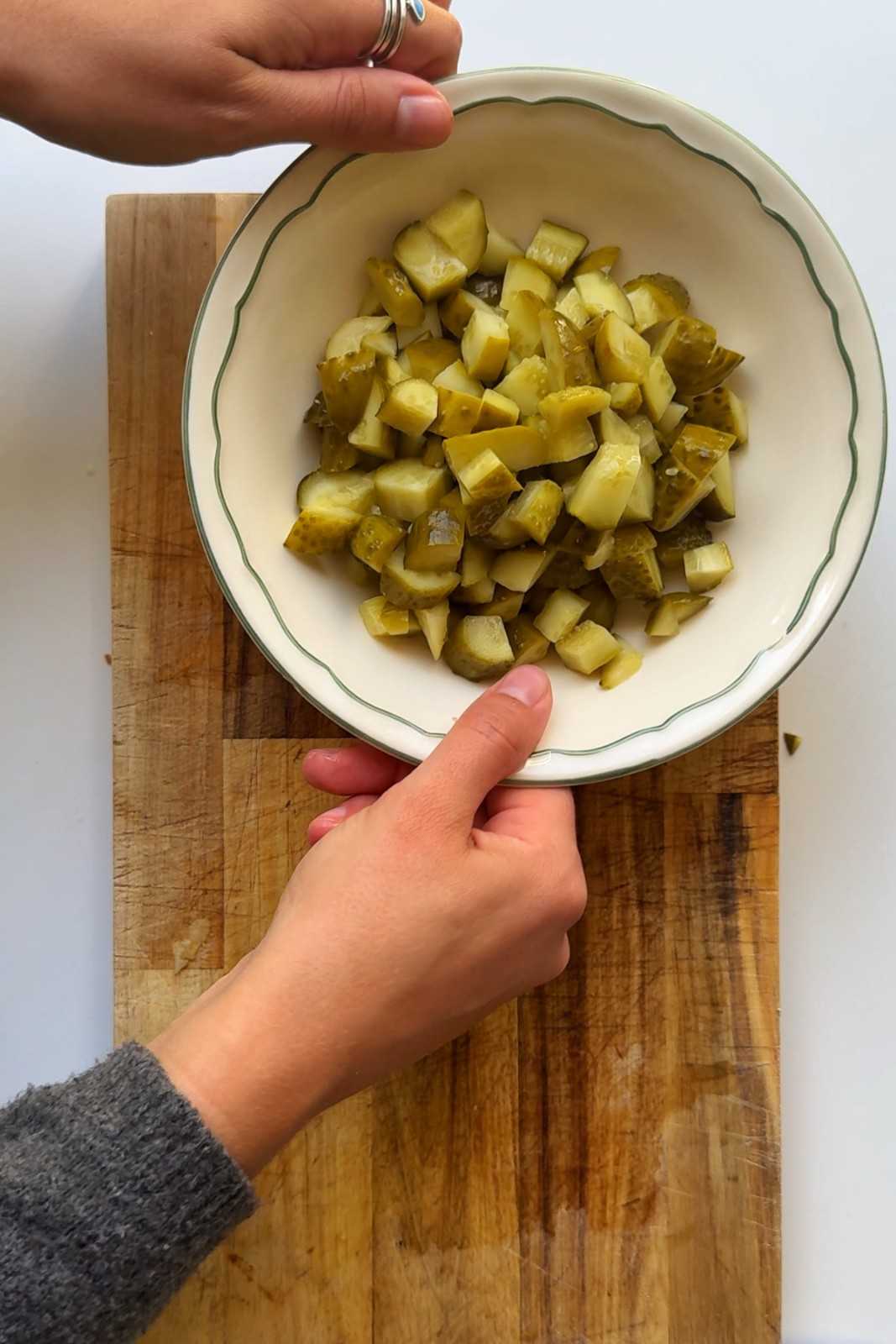 Hands holding a white ceramic bowl of chopped dill pickles on a wooden cutting board.