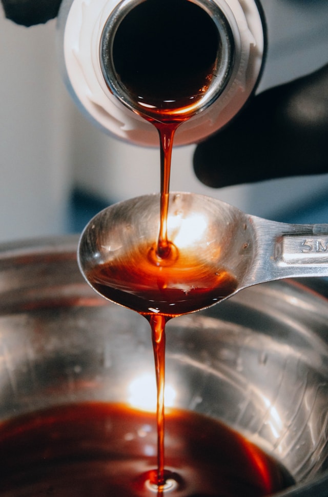 Dark corn syrup being poured from a bottle onto a metal measuring spoon over a mixing bowl.