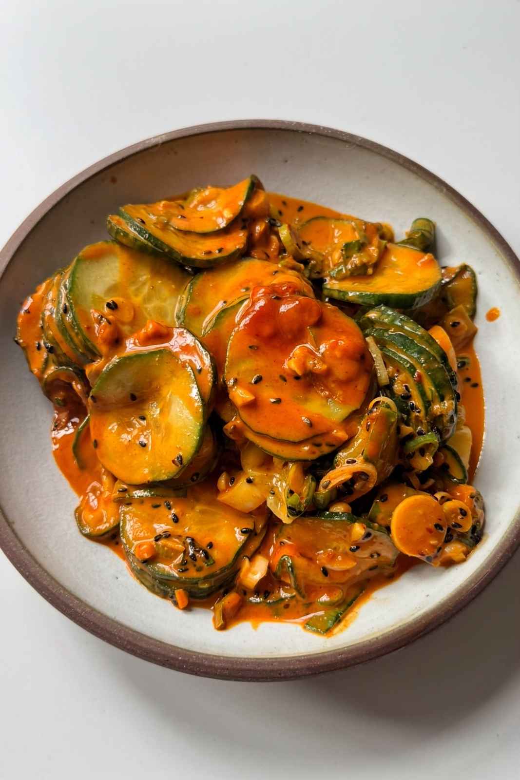 A white bowl of spicy cucumber salad with thinly sliced cucumber rounds glossy with red gochujang peanut dressing, finely sliced spring onions, white sesame seeds, and crispy chili oil drizzled over the top.