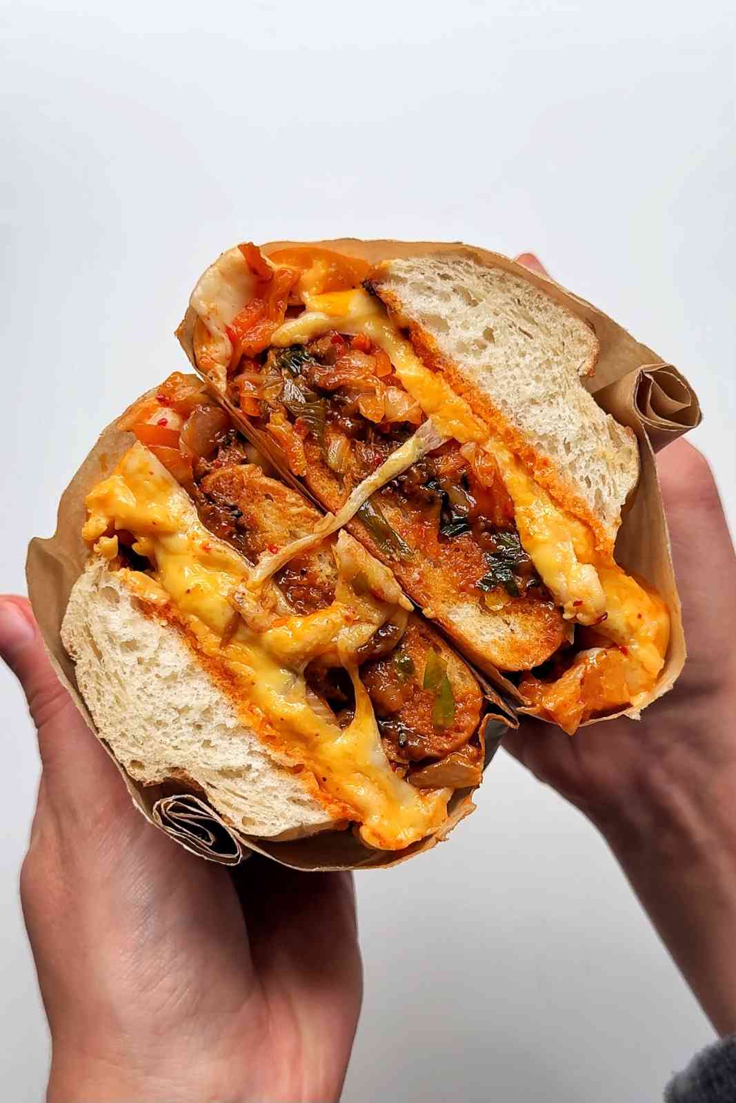 A bulgogi cheesesteak sandwich wrapped in brown parchment paper, sliced in half and stacked to show layers of thinly sliced marinated Korean beef, kimchi, spring onions, and melted American and mozzarella cheese oozing out of a white bread roll.