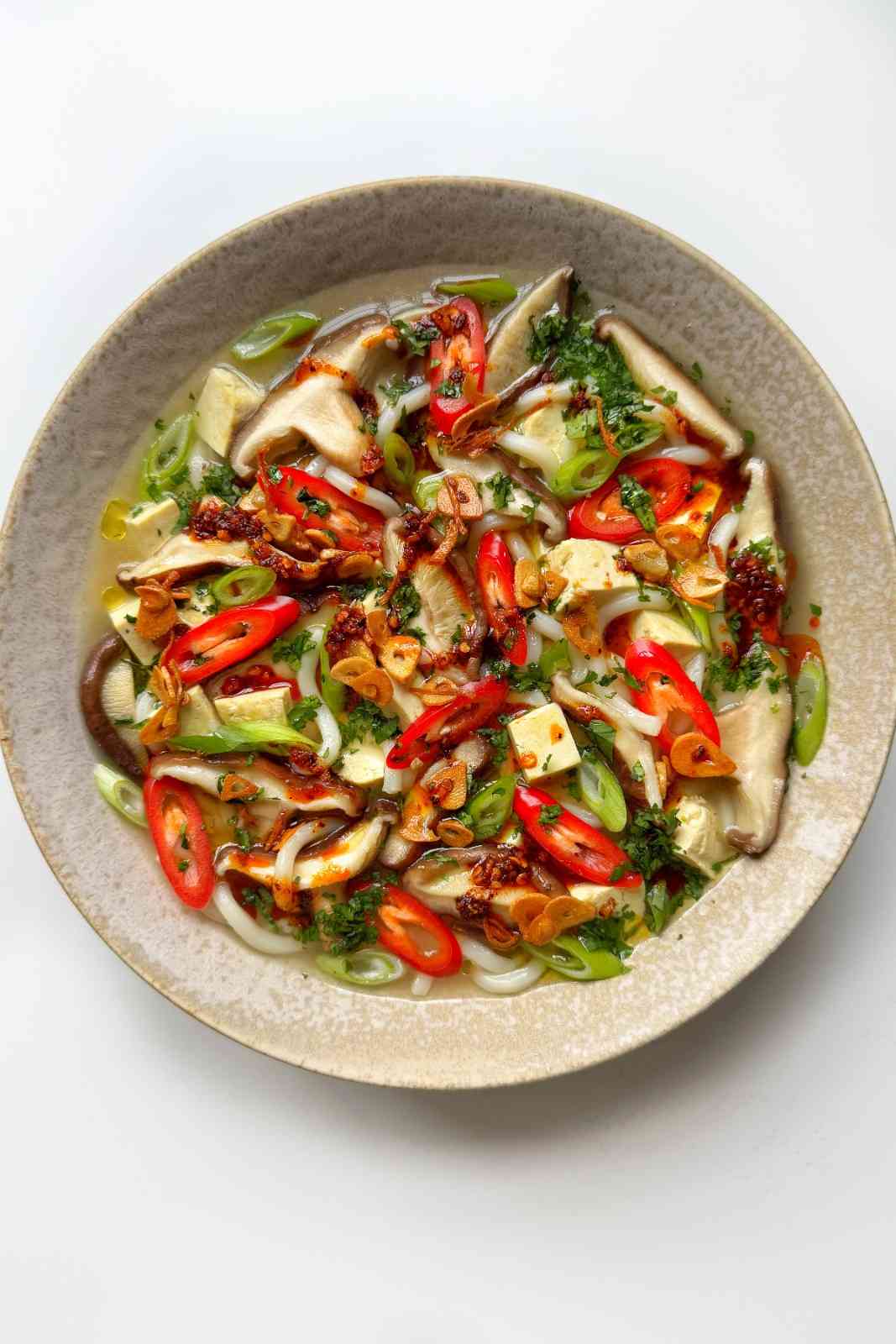 Overhead bowl of miso soup with mushroom, udon noodles, and tofu cubes in a clear broth, topped with sliced red chili, scallions, cilantro, and crispy garlic ginger oil.