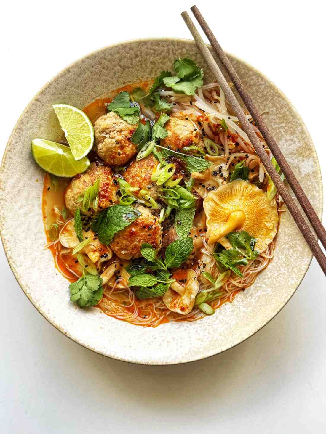 Overhead bowl of Thai-style pork meatball noodle soup with golden seared meatballs, rice noodles, mushrooms, and bean sprouts in a spicy broth, topped with cilantro, fresh mint, scallions, and lime wedges.
