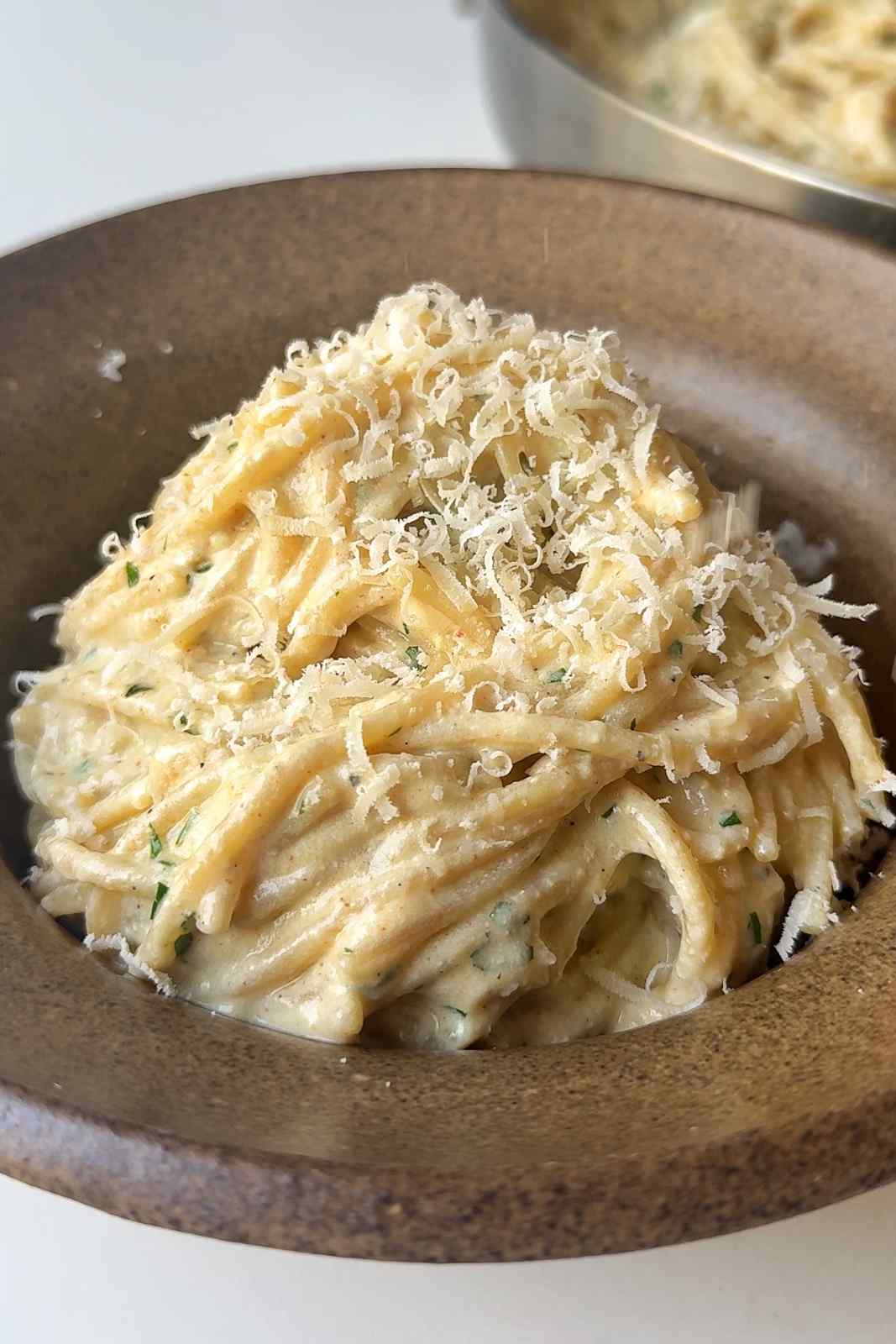 Creamy silken tofu pasta sauce coated over a swirl of spaghetti, topped with a mountain of finely grated Parmesan cheese in a brown bowl.