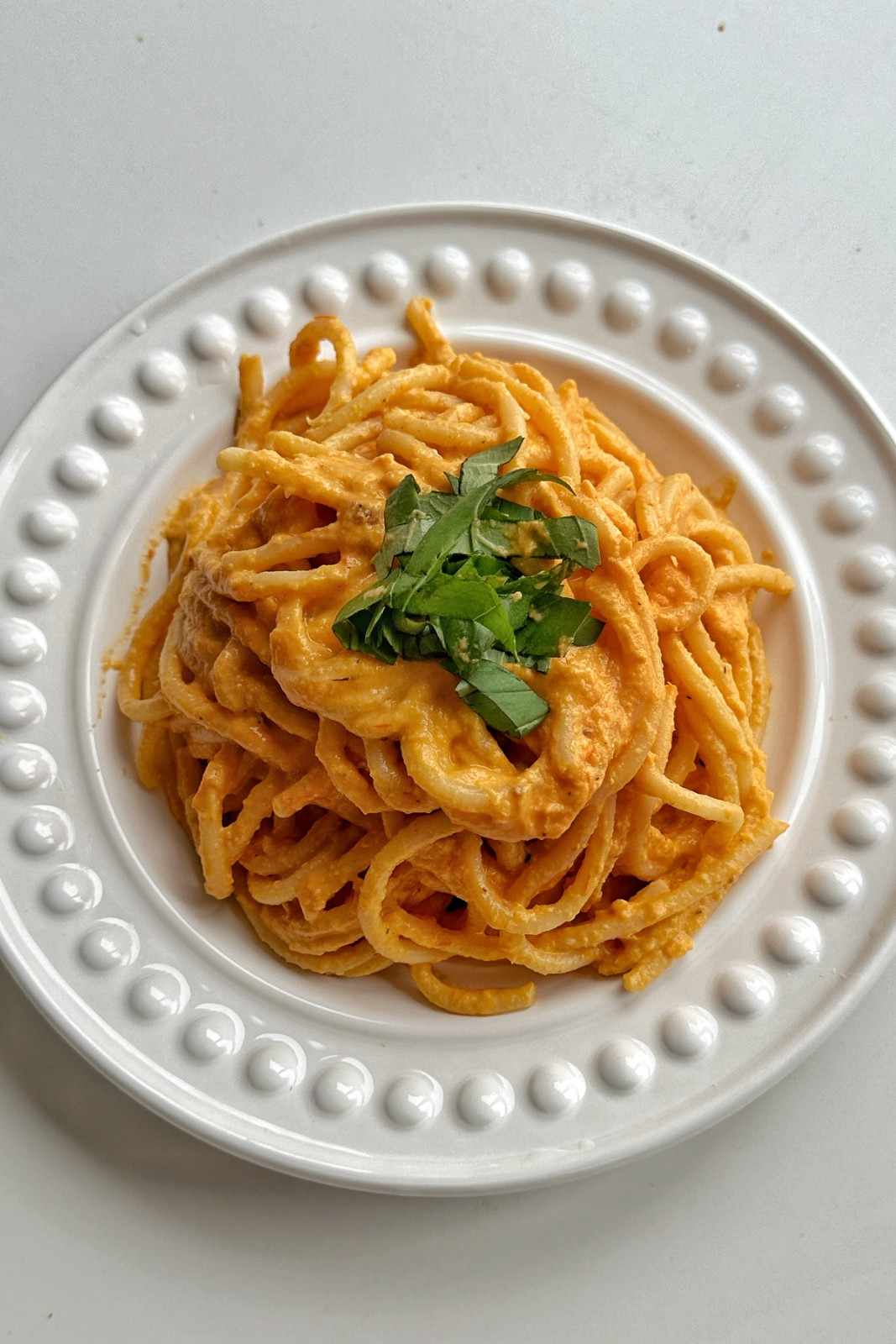 A top-down view of thick bucatini pasta tossed in a creamy, rich orange-pink roasted tomato and feta sauce, served on a white plate with a raised beaded rim and topped with shredded fresh basil.
