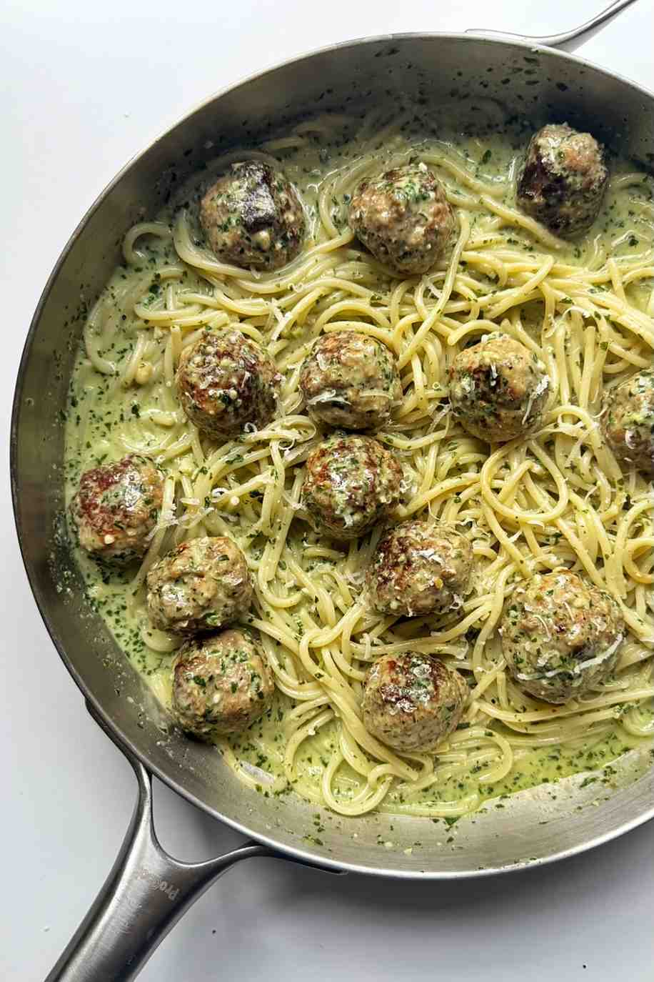 An overhead shot of a large stainless steel skillet filled with spaghetti tossed in a creamy, light-green pesto sauce, dotted with several golden-brown homemade meatballs and sprinkled with grated cheese.
