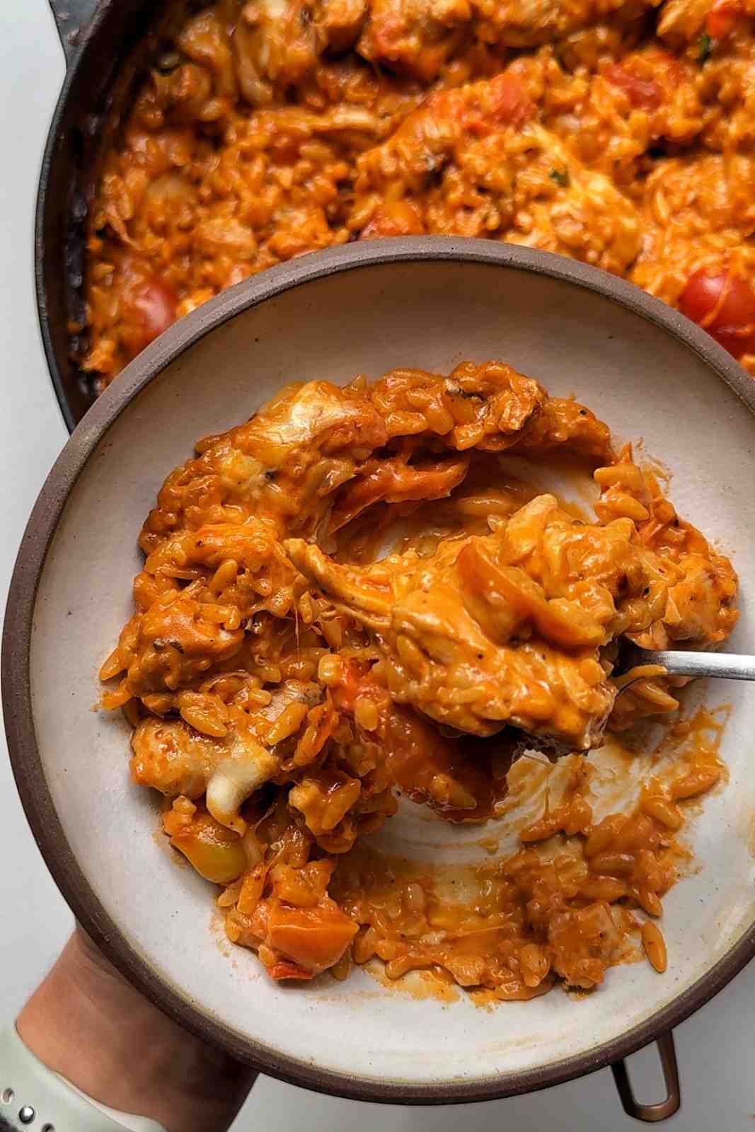 A close-up shot of a ceramic bowl being filled with creamy, orange-red chicken orzo, showing melted mozzarella cheese stretching from the spoon, with the larger cooking pan visible in the background.