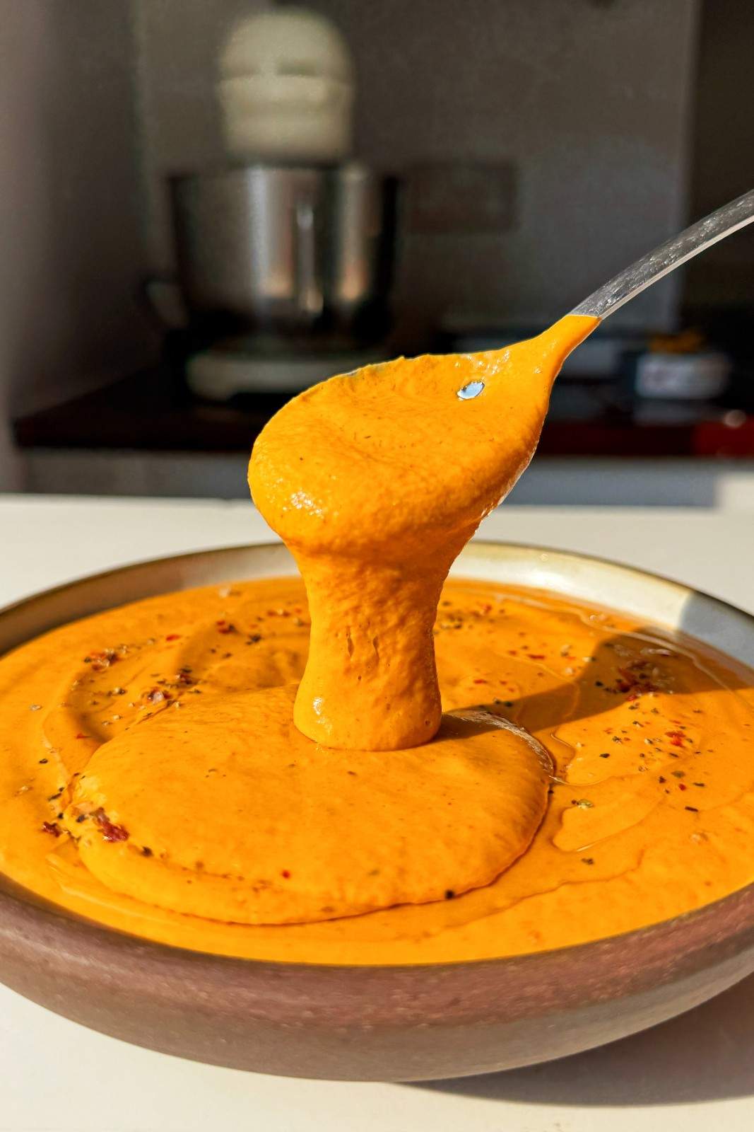 Smooth, creamy roasted red pepper pasta sauce being spooned from a dark bowl, flecked with chili flakes and black pepper.
