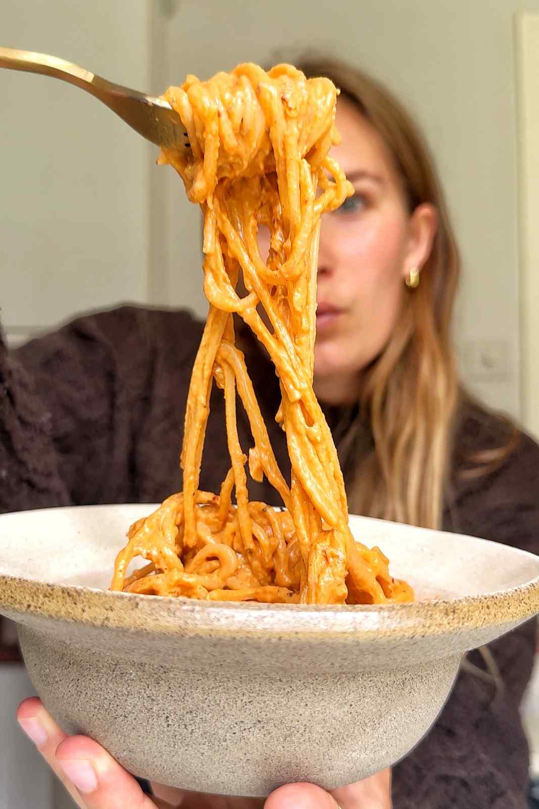 A close-up shot of a large forkful of spaghetti being lifted out of a rustic beige ceramic bowl, showing thick strands of pasta coated in a creamy, orange sun-dried tomato sauce with Emily blurred in the background.