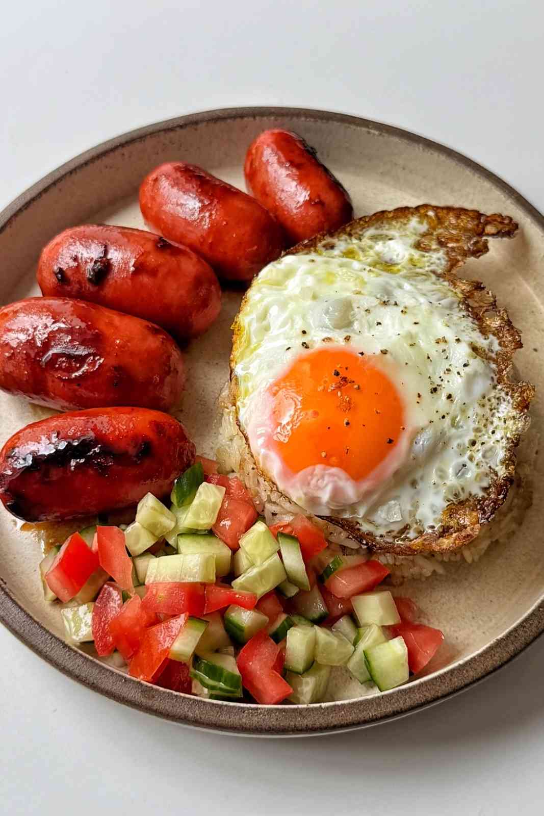silog breakfast with fried egg, cucumber and tomato, and sausages on a plate.