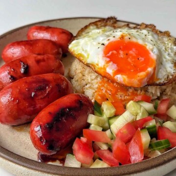 a plate of sausages, garlic fried rice, fried eggs, tomatoes, and cucumbers.